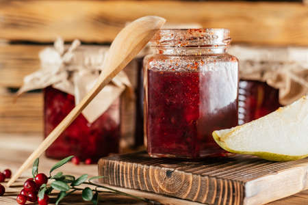 Jars of homemade lingonberry and pear jam with craft paper on lids on wooden surface next to fresh lingonberries and pears.の写真素材