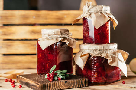 Jars of homemade lingonberry and pear jam with craft paper on lids on wooden surface next to fresh lingonberriesの写真素材