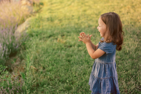 Little girl 3-4 with dark hair in denim dress claps and laughs standing on green lawnの写真素材