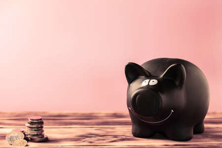 Black piggy bank and coins on a wooden table on a pink background, copy space.の写真素材