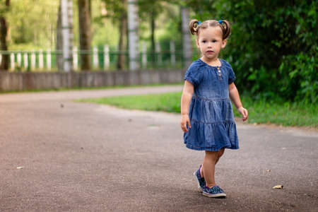 Little smiling cute dark-haired girl 1-3 in a denim dress walks along the road in the park against the backdrop of greenery.の写真素材