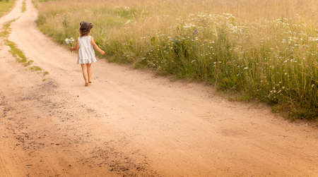 Little cute girl 1-3 in a light dress, running on a path in the field on a background of grass with a bouquet of daisies in summerの写真素材