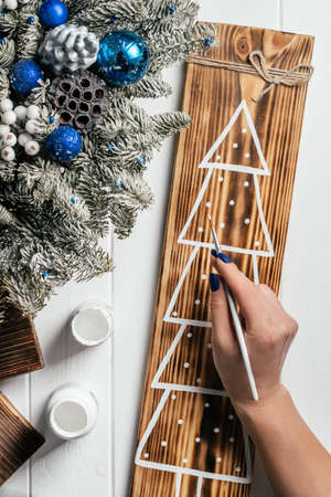 Womans hand draws Christmas tree on decorative board made of burnt wood with brush of white paint....