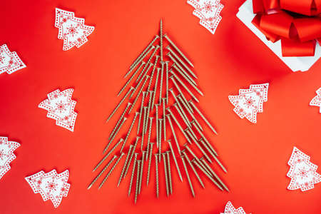 Long golden self-tapping screws, laid out in shape of Christmas tree, white wooden Christmas tree decorations and gift box on red backgroundの写真素材