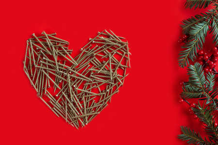 Long golden self-tapping screws, laid out in shape of heart with Christmas tree branches and red berries on red backgroundの写真素材
