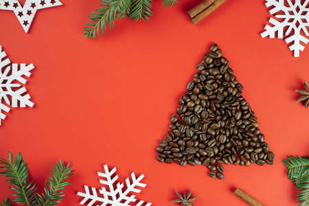 Brown coffee beans laid out in shape of Christmas tree with spruce branches and wooden snowflakes on red background. Flat lay. Copy spaceの写真素材