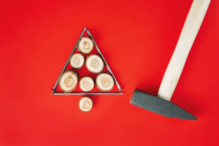 Long nails and wood cuts or branch slabs, laid out in shape of Christmas tree with hammer on red background. Flat layの写真素材
