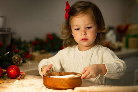 Cute little serious girl 2-4 with a red bow prepares Christmas gingerbread cookies in the New Year's kitchen.の写真素材