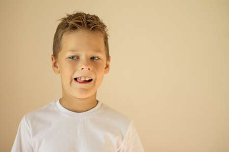 A 7-10 boy in a white T-shirt is wobbling a falling tooth with his tongue on a beige background, copy spaceの写真素材