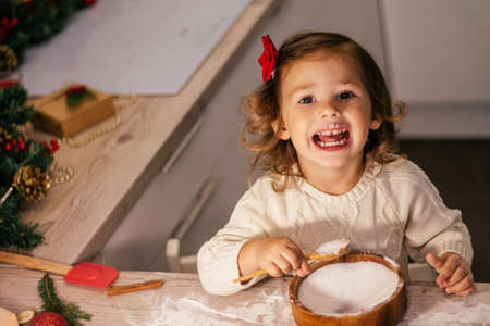 Cute little smiling girl 2-4 with a red bow prepares Christmas gingerbread cookies in the New Year's kitchen.の写真素材