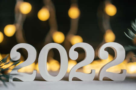 Happy New Year 2022. White wooden numbers 2022 stand on white surface with Christmas tree branches against background of garland light bulbs. Merry Christmas. Copy spaceの写真素材