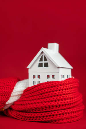 White toy house wrapped in red scarf on red background. Heating or insulating buildings or houses concept. Copy space. Verticalの写真素材