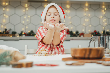 Little dark-haired girl 3 years old in red Christmas cap and checkered shirt prepares dough for gingerbread cookies in white Christmas decorated kitchen with garland lights. Merry Christmas, New Yearの写真素材