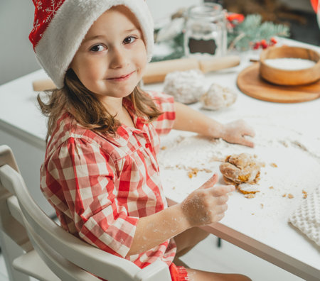 Little dark-haired girl 3 years old in red Christmas cap and checkered shirt prepares dough for gingerbread cookies in white Christmas decorated kitchen with garland lights. Merry Christmas, New Yearの写真素材