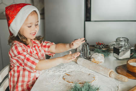 Little dark-haired girl 3 years old in red Christmas cap and checkered shirt prepares dough for gingerbread cookies in white Christmas decorated kitchen with garland lights. Merry Christmas, New Yearの写真素材