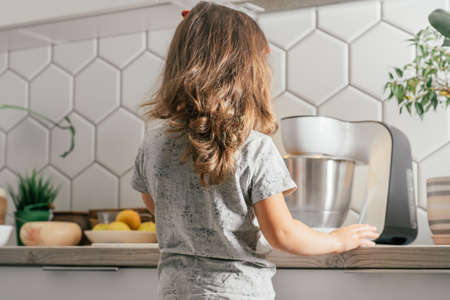 Little dark-haired girl 3 years old in red headband bakes apple pie in kitchen. Child stands near planetary mixer. Children help on household chores. Kid cooking food. Back viewの写真素材
