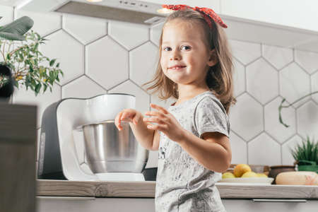 Little dark-haired girl 3 years old in red headband bakes apple pie in kitchen. Child stands near planetary mixer and smiles. Children help on household chores. Kid cooking foodの写真素材