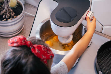 Little dark-haired girl 3 years old in red headband bakes apple pie in kitchen. Child turns on planetary mixer. Children help on household chores. Kid cooking food. Top viewの写真素材
