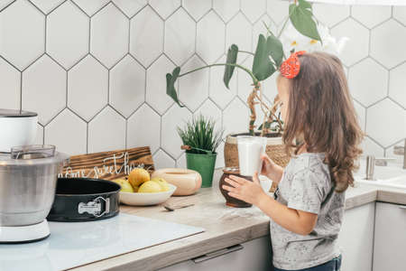 Little dark-haired girl 3 years old in red headband bakes apple pie in kitchen. Child holds measuring cup with flour. Children help on household chores. Kid cooking foodの写真素材