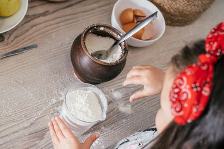 Hands of little girl in red headband bake apple pie in kitchen. Child puts flour in measuring cup. Children help on household chores. Kid cooking food. Top viewの写真素材