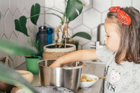 Little dark-haired girl 3 years old in red headband bakes apple pie in kitchen. Child adds apples to dough. Children help on household chores. Kid cooking foodの写真素材