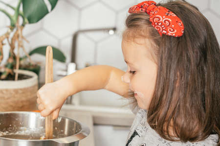 Little dark-haired girl 3 years old in red headband bakes apple pie in kitchen. Child mixes dough in bowl. Children help on household chores. Kid cooking foodの写真素材