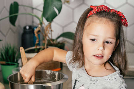 Little dark-haired girl 3 years old in red headband bakes apple pie in kitchen. Child mixes dough in bowl. Children help on household chores. Kid cooking foodの写真素材