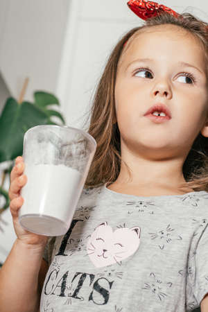 Little dark-haired girl 3 years old in red headband bakes apple pie in kitchen. Child holds measuring cup with flour. Children help on household chores. Kid cooking food. Verticalの写真素材