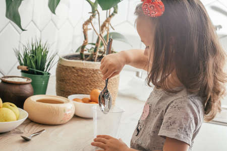 Little dark-haired girl 3 years old in red headband bakes apple pie in kitchen. Child puts sugar in measuring cup. Children help on household chores. Kid cooking foodの写真素材