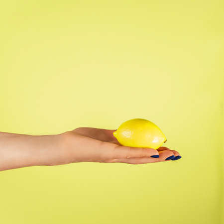 Female hand with blue nails holding fresh ripe juicy lemon isolated on yellow. Squeeze out juice. Diet topic. Creative art concept. Food minimal colorful concept. Vertical. Copy space. Mock up.の写真素材