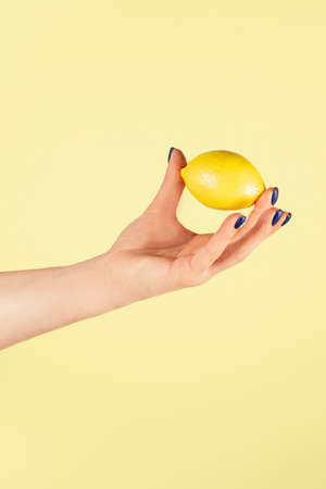 Female hand with blue nails holding slightly with two fingers fresh ripe juicy lemon isolated on yellow. Creative art concept. Food minimal colorful concept. Vertical. Copy space. Mock up.の写真素材