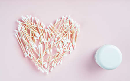 Wooden cotton buds laid out randomly in shape of heart and white cosmetic jar on pink background. Auricle hygiene. Beauty concept. Heart relation concept. Flat lay. Copy space. Mock up.の写真素材