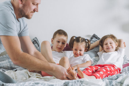 Father plays with three children on bed. Dad tickles kids feet. Family of daddy, two girls and boyの写真素材
