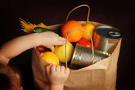 Little girl puts products in a paper bag on a brown background. A child takes food out of a food delivery service package. Canned food, pasta, vegetables, fruits. Contactless delivery.の写真素材