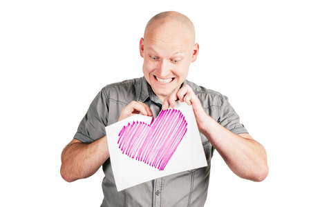 Smiling handsome bald middle-aged man in gray shirt tear pink heart drawn on sheet of paper with pleasure isolated on white background. Valentines day, hatred, dislike, relations, emotion conceptの写真素材