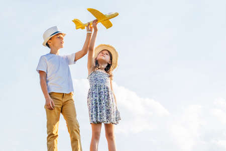 Portrait of children boy and girl playing outdoors. Light sunny day. Cheerful and calm concept. Fresh air in countryside. Kids wearing hats and playing flying yellow toy airplane. Planning travellingの写真素材