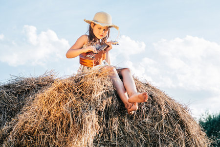 Portrait of little girl playing small guitar ukulele, plunking on haystack in field. Light sunny cloudy day. Cheerful and enjoy concept. Trees on background. Wearing dress and hat. Concentrated kidの写真素材