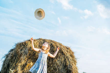 Portrait of little girl in blue dress catching hat, standing near haystack in field. Light sunny cloudy day. Joy and glad concept. Blue clear sky on background. Wind taking away straw hat. Low angleの写真素材