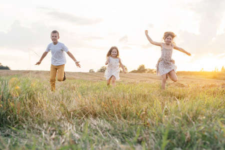 Portrait of three children playing game of catch, jumping and running on dry grass hay field paths in sunset. Trees and meadow on background. Looking around. Cloudy sunny sky. Haying timeの写真素材