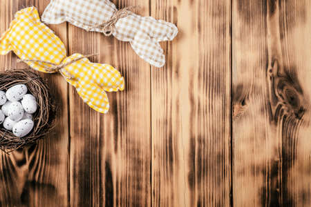 Composition of two handmade square brown, white and yellow colored bunnies with string-scarf on wooden natural background and white spot quail eggs in bird nest. Celebrate Easter holiday. Copy spaceの写真素材