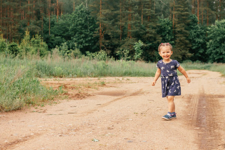 Beautiful happy little girl 2-3 years old in blue dress and sandals running along country road, in summer field near forest on warm sunny day.の写真素材