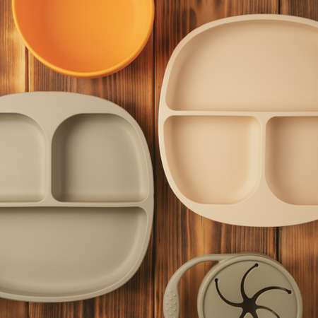 Composition of modern colorful pastel silicone food trays, bowl and snack cup on brown wooden table. Baby tableware, first feeding, serving concept.の写真素材