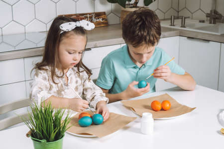 Portrait of studiously two little children, boy and girl, painting brush multi coloring Easter hens eggs on paper plate table, plant in kitchen. Making white art patterns. Traditional holiday event.の写真素材