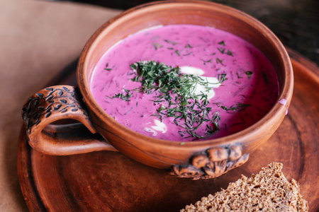Delicious tasty thick purple pink beetroot soup on kefir cultured buttermilk in earthenware clay carved pattern. Tureen with handle on plate with black rye bread and green dill. Lunch time. Close upの写真素材