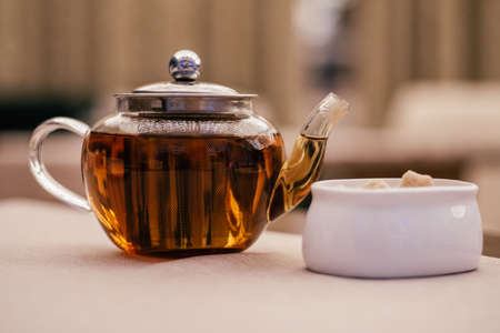 Freshly brewed strong yellow brown tea in filling transparent glass teapot with metal lid, white...