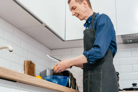 Portrait of middle-aged man wearing blue long-sleeve shirt and black apron breaking egg with knife...
