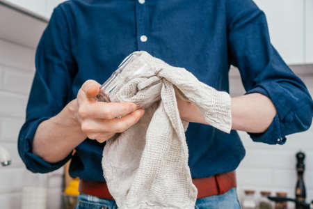 Close up of unrecognizable man in blue shirt, jeans and red leather belt drying glass using grey cotton kitchen towel standing in bright white modern kitchen. Housework, cleaning concept.の写真素材