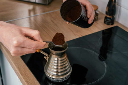 Close up of hands of unrecognizable man, putting full heaped tea spoon of natural ground coffee from round glossy tin in copper cezve on black electric cooker for brewing near spice grinder in kitchenの写真素材