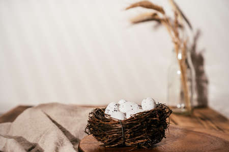 Easter composition of quail spot eggs in nest on wooden table and branch of wheat in glass on beige...