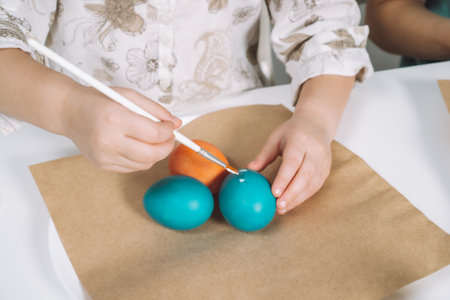 Cropped portrait of girl hand, painting multi coloring Easter hens eggs on brown paper plate table with paintbrush. Making art design white patterns on blue, red animal eggs. Holiday event. Close upの写真素材